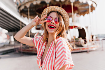 Outdoor portrait of stunning girl posing with peace sign near carousel. Female model with pleased smile dancing in amusement park.