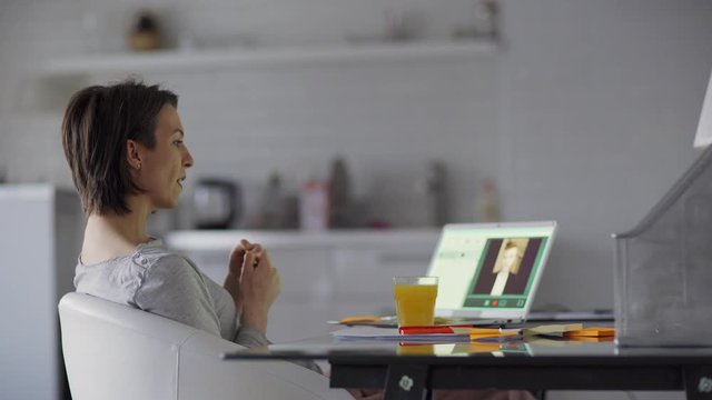 Side view medium shot of confident middle aged woman having business video call on laptop computer at home. Businesswoman drinking orange juice and talking to male partner