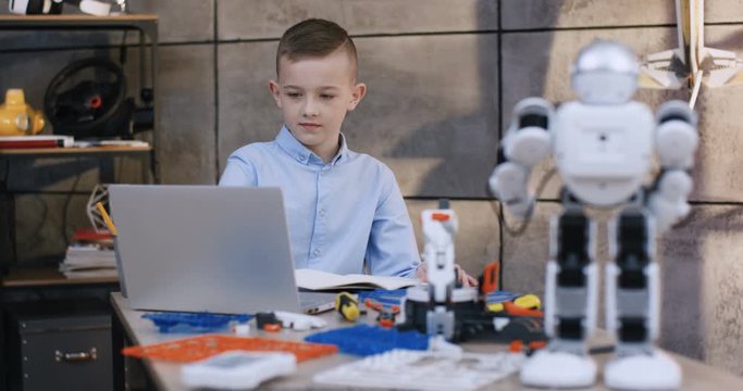 Nice and smart teen Caucasian boy in the blue shirt working at the laptop computer at the desk while constructing some model from constructor for kids. Toy robot on the first plan.