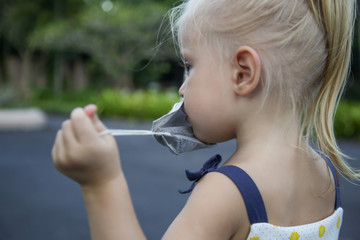 Little toddler girl trying to put medical protective mask. Candid outdoor portrait of child with medical mask. Corona virus outbreak or air pollution concept.	