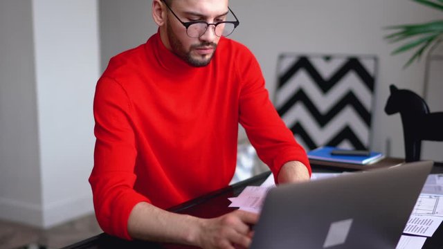 Young businessman keyboarding information on laptop to check online database sitting at table in modern apartment.Male entrepreneur in casual wear working remotely at home interior
