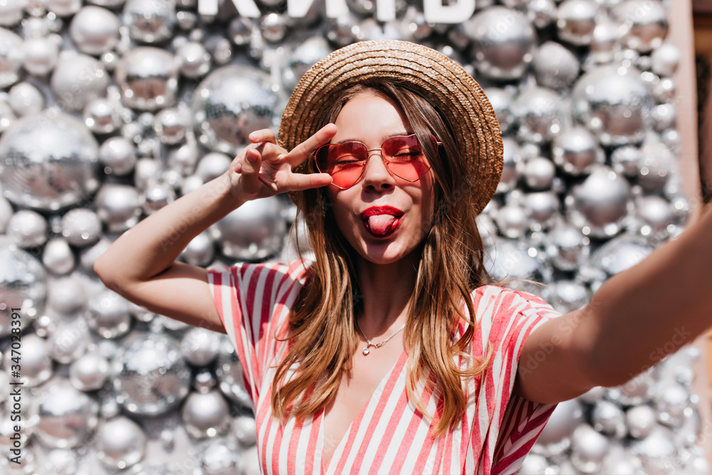 Wall mural Stunning young woman in pink sunglasses making selfie on sparkle background. Blithesome girl in straw hat posing with tongue out near disco balls.