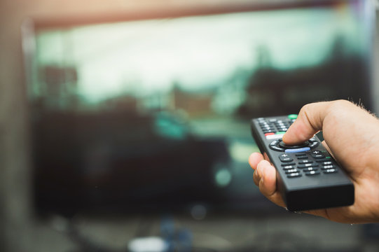 Young Man Holding Television Remote Control. Hands Pointing To Tv Screen Set And Turning It On Or Off Select Channel Watching Tv On His Sofa At Home In The Living Room Relax..