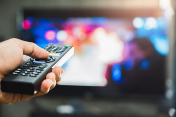 Young man holding television remote control. Hands pointing to tv screen set and turning it on or off select channel watching tv on his sofa at home in the living room relax.. - obrazy, fototapety, plakaty