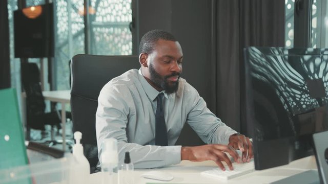 Tired African American handsome man uses computer for work in modern office programmer serious looks at monitor screen workplace engineering problems slow motion