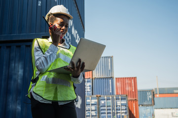 Black foreman woman worker working checking at Container cargo harbor holding laptop computer and walkie-talkie to loading containers. African dock female Logistics import export shipping concept.