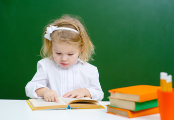 Little girl student reads a book while sitting against a green school board