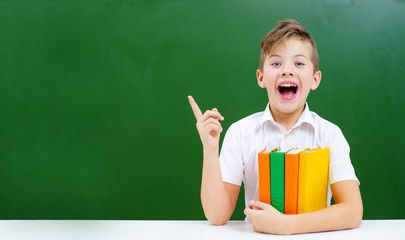 The boy sitting on the background of the school blackboard hugs a stack of books and with a joyful smile shows his finger towards the empty space on the blackboard