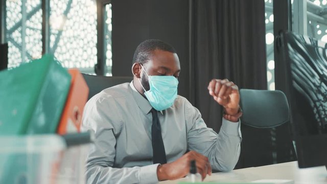 Tired African American man wearing mask feel headache uses computer for work in modern office programmer serious looks at monitor screen businessman coronavirus close up slow motion