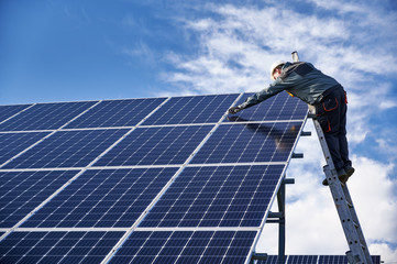 Male worker in white safety helmet standing on ladder and repairing photovoltaic solar panel...