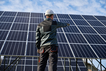 Low angle view on technician in dark blue uniform standing with his back to the camera, pointing at solar plant. Rear view. Alternative ecological source of energy concept