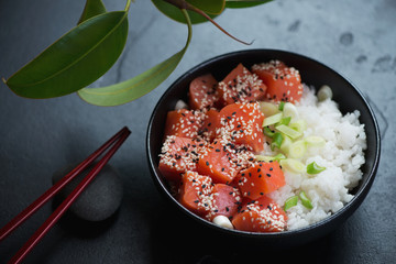 Poke bowl with salmon fillet, white rice, green onion and sesame over black stone background, studio shot
