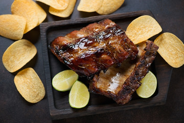 Wooden serving tray with barbecued pork ribs and chips, studio shot on a dark brown metal background, elevated view