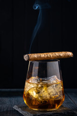 A glass of whiskey or cognac with ice cubes and a Smoking cigar lying on top, black wooden...