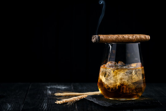 A glass of whiskey or cognac with ice cubes and a Smoking cigar lying on top, black wooden background