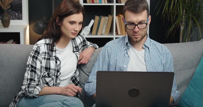 Happy young couple in casual clothing sitting on sofa at home and using portable computer during free time. Concept of quarantine and relationship