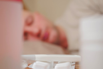 Obraz na płótnie Canvas White thermometer on a pack of pills and two bottles with medicine in focus, sick man sleeping in bad out of focus in the background.