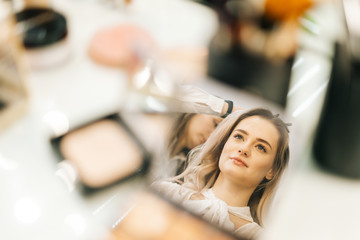 Close-up of face of young woman reflected in mirror of powder on woman table, looking away. Female hairdresser making hairstyle in beauty salon. Concept of backstage work.