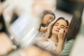 Close-up of face of young woman reflected in mirror of powder on woman table. Professional stylist and makeup artist doing makeup and style hair for young woman. Concept of backstage work.