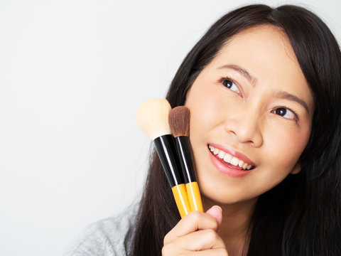 Asian woman has dark brown long hair, is makeup artist, holding brush and make up cheek bone aspink nude tone. Lady is smiling, happy and cheerful to be beautiful stylelist concept.  Female see above.
