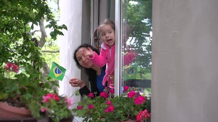 Covid-19. Brazilian family mother and little girl child on balcony / window are smiling and waving the flag of Brazil. Self-isolation in quarantine, lockdown, stay at home, social distancing - Powered by Adobe