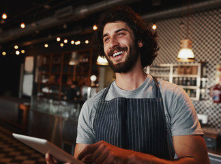 Handsome caucasian coffee shop owner laughing while holding digital tablet in hand