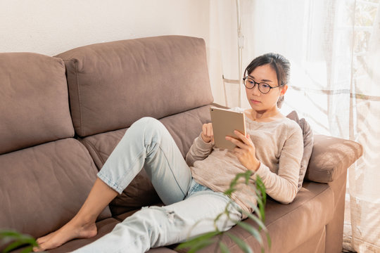 Millennial Asian Woman With Glasses Lying On The Sofa Watching Ipad Tablet. Girl At Home Laying On The Couch Watching Videos. People Enjoying Indoors. Quarantine At Home.