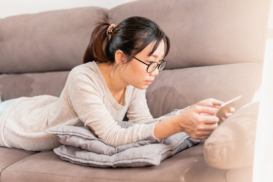 Young Asian Mother Enjoying Her Time Alone Lying On The Sofa Watching Ipad Tablet. Girl Enjoying At Home On The Couch Watching Movies.