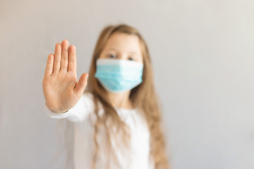 A cute serious girl of European appearance stands with a medical mask on her face and shows a sign, tin stop with her hand. Hand in focus, the girl in defocus. Place for text