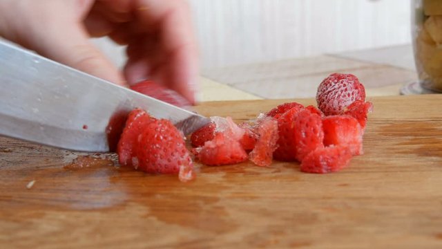 Female hands cut a frozen strawberry with a knife for future fruit salad or smoothie on a wooden kitchen board. Morning breakfast at home.