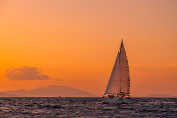 Obraz na płótnie Canvas Sailing boat floating on the blue Aegean Sea during sunrise / sunset with open main sail and genoa. Orange sky with clouds and islands in background for copy space
