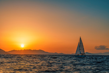 One sailing boat floating on the blue Aegean Sea during sunrise / sunset with open main sail and...