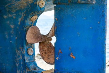 An old scrappy and rusty propeller screwed at the rear of a blue ship hull. Rough blue surface with...