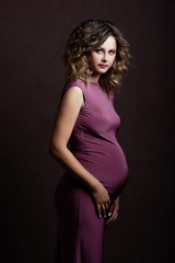 A young pregnant woman looks forward, holds her hands under her stomach, beautiful makeup and hairstyle, behind her brown background
