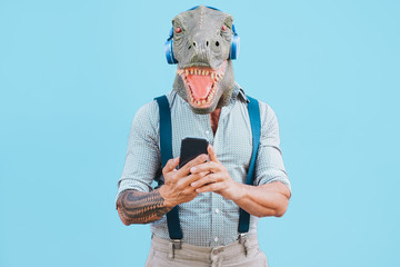 Senior tattooed man with t-rex mask using smart mobile phone while listening music during...