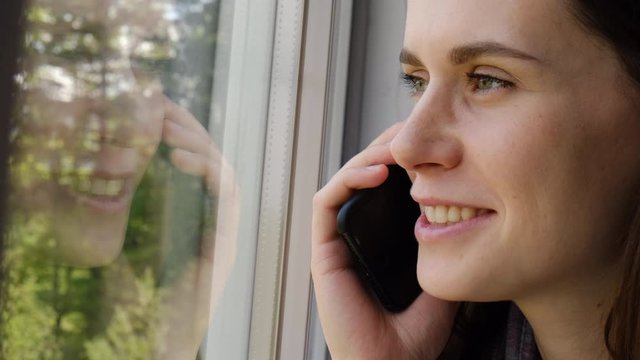 Joyful beautiful young woman talking on smartphone at home, smiling cute girl making answering call by cellphone, beautiful lady having pleasant funny conversation speaking by mobile sitting on sill