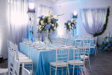 the restaurant has beautiful tables with serving, the decor is made in blue, the tables are indicated with flowers and beautiful dishes, a wedding celebration, there are white chairs for guests