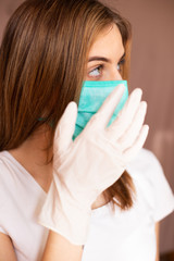 portrait of beautiful young woman wearing cotton green mask and white medical/surgical gloves on pastel background. She is adjusting mask. 