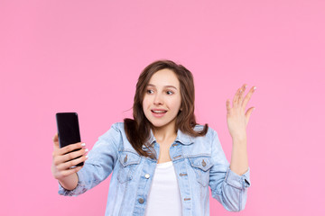 Pretty, surprised, shocked, .indignant girl is holding in hand smartphone, looking at screen display. Young woman dressed in denim jacket and white t-shirt on pink background. Modern devices concept.