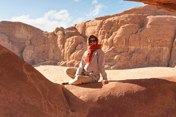 Young woman wearing warm jacket and scarf around head smiling, sitting at naturally formed rock window in wadi rum desert on sunny day