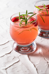 Refreshing summer cranberry cocktail with ice and rosemary on white concrete background close up.