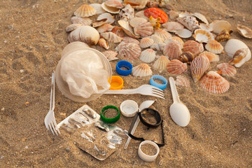 A lot of different plastic waste: disposable plastic cups,plastic bags, disposable spoons, forks, plastic lids, collected on the beach against the background of sand and beautiful shells. 