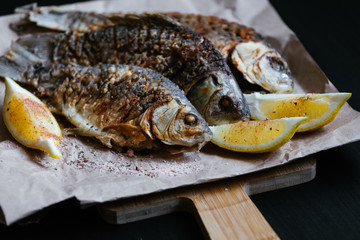 National dish of river fish. Grilled carp, crucian carp with lemon slices and salt on a wooden board and craft paper on a black wood background
