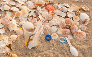 A lot of different plastic waste: disposable plastic cups,plastic bags, disposable spoons, forks, plastic lids, collected on the beach against the background of sand and beautiful shells. 