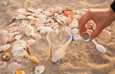 A man's hand collects and removes plastic waste on the beach: bags, disposable plastic appliances, plastic lids among the shells. The concept of ecology, cleaning the environment
