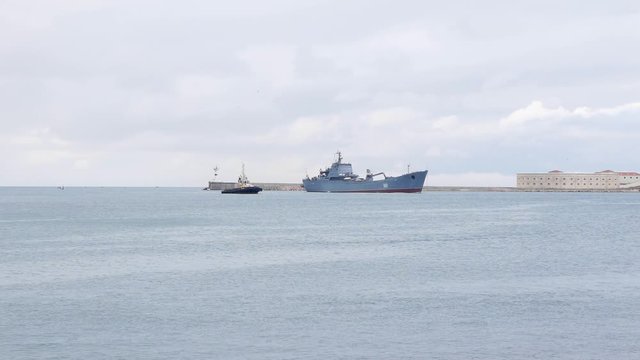 A warship accompanied by a tugboat enters the bay. Overcast sky