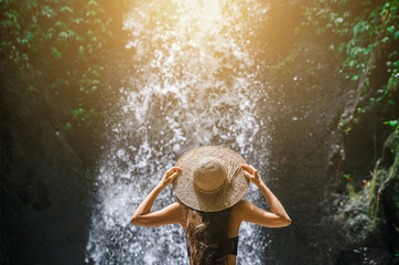 Beautiful young woman tourist with straw hat from back in the tropical jungle looks at a strong and...