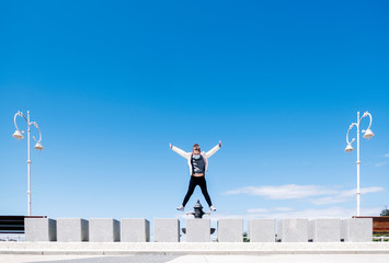 A girl wearing a safety mask has fun jumping through a concrete wall from a walk with streetlights in the city with blue sky on a sunny day. Copy space.