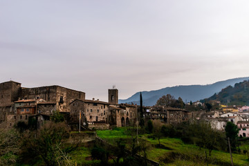 Fototapeta na wymiar A panoramic view of Santa Pau (Girona, Spain), its old town, fortress, and other building, with the Pyrenees mountains in the background, in the cloudy afternoon in winter