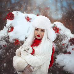 A beautiful sweet girl in a hat with earflaps with a white cat in her hands, stands in winter near a Rowan Bush. Russian winter.